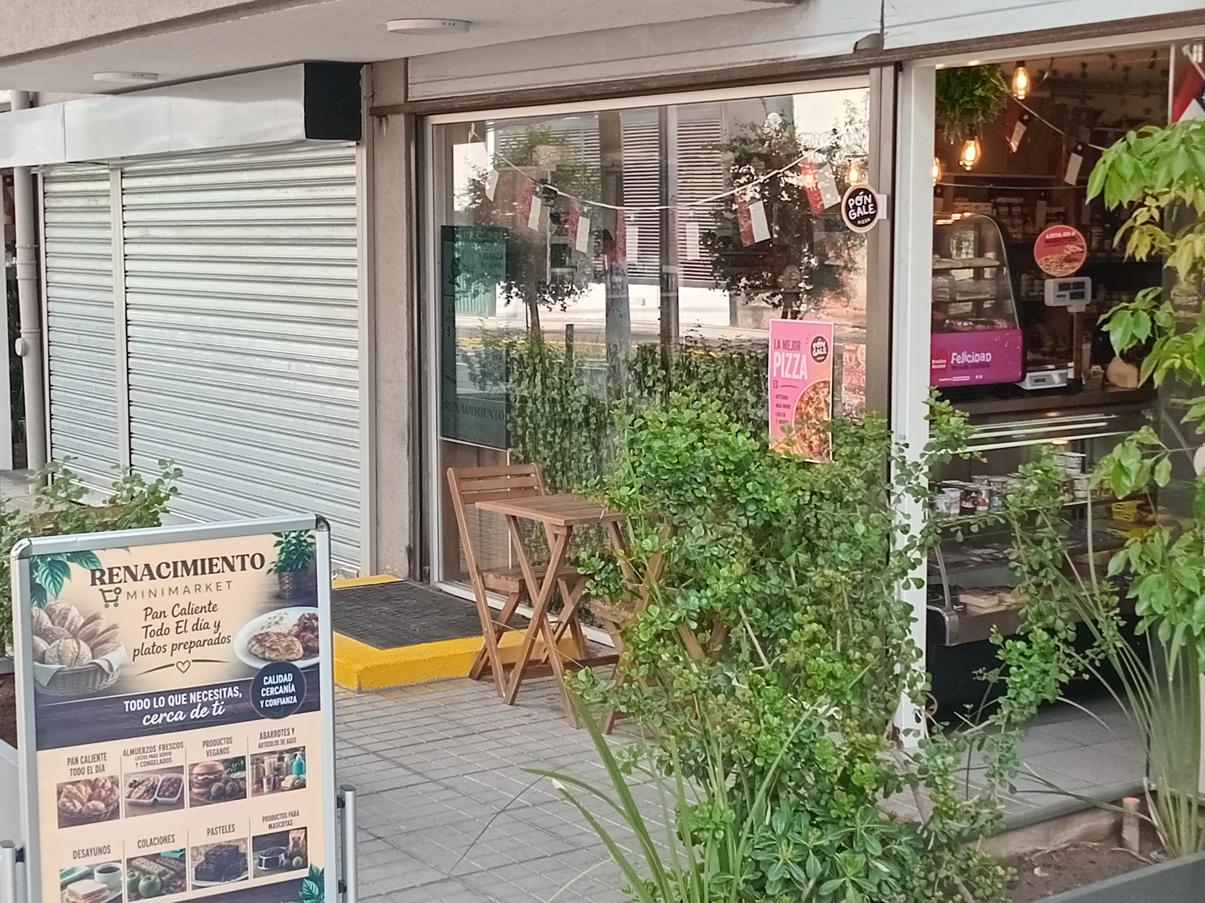 Fachada de Renacimiento Minimarket, con su letrero en la vereda, mesa de madera y plantas
